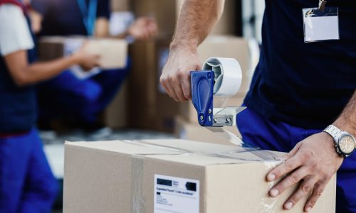 Close-up of delivery man closing carboard box with a tape while preparing packages for shipment.
